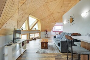 Een loft-achtige kamer met een schuine kap, een eethoek met een tafel en stoelen, een bank en een televisie op een witte standaard.