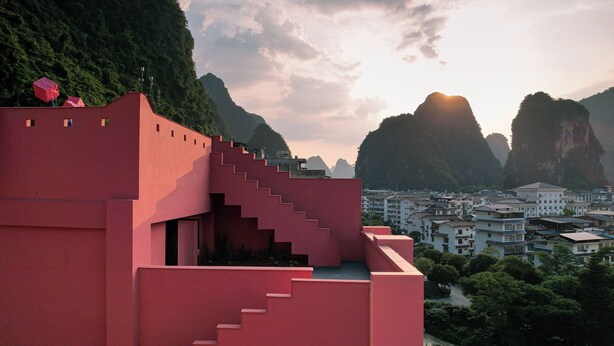 A red building with a staircase leading to a flat roof, surrounded by a mountainous landscape and a town below. - Yangshuo B&G Hotel (Guilin)