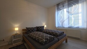 A bedroom with a bed, two bedside tables, a window with blinds, and a radiator.