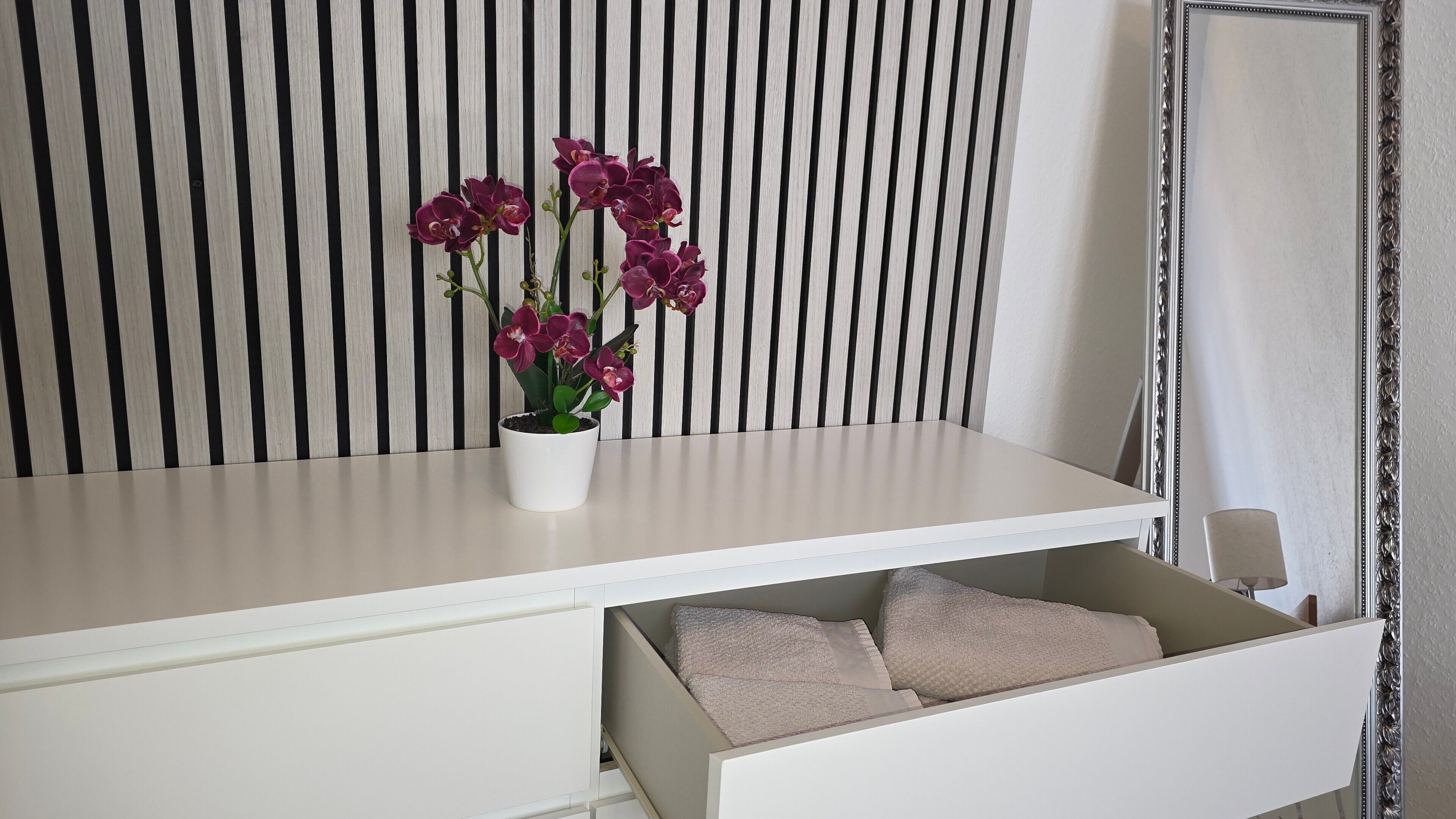 Une table de chevet blanche, ornée d’une orchidée en pot et dotée d’un tiroir contenant des coussins de couleur beige.