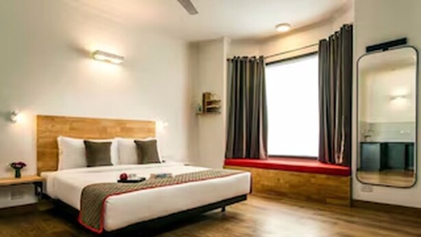 Una habitación de hotel moderna con suelo de madera, una cama grande con cabecera, una ventana con cortinas y un espejo en la pared.