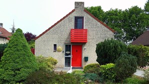 Une maison avec une porte rouge, un petit balcon et un jardin agrémenté de divers arbustes.