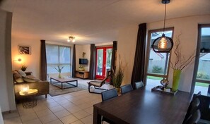 Een moderne woonkamer met een bank, salontafel, eettafel en stoelen. Er is een televisie op een standaard, een rode deur en grote ramen.