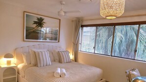 Un dormitorio con cama, mesita de noche, lámpara, un cuadro y ventana con persianas.