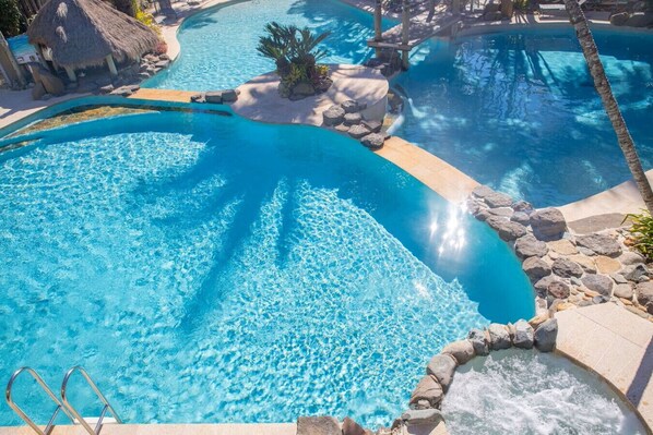 Una piscina de varios niveles con agua azul cristalina, rodeada de rocas y palmeras, e incluye un área de hidromasaje.