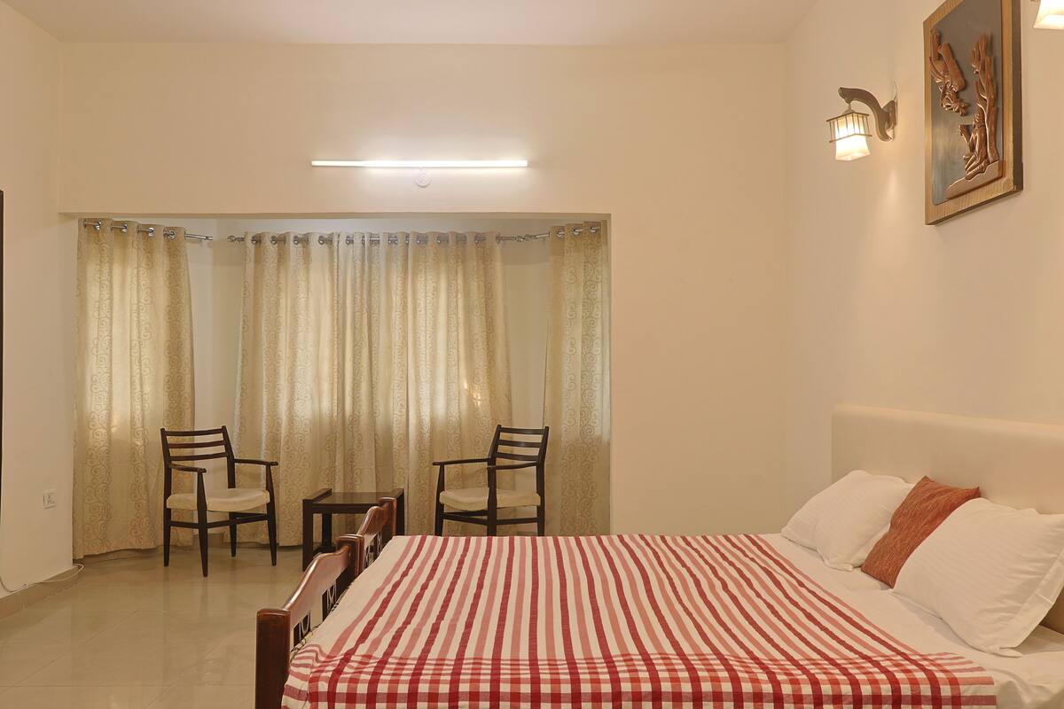 A bedroom with a bed, two chairs, a table, a window with curtains, a wall-mounted light, and a painting.