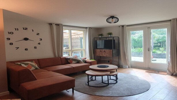A modern living room with a sectional sofa, a round coffee table, a wall clock, a television, and large windows. - Forest Bungalow w/ Sauna for 6 (Ewijk)