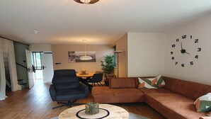 A living room with a sectional sofa, a round coffee table, a dining area with chairs, and a wall clock. - Forest Bungalow w/ Sauna for 6 (Ewijk)