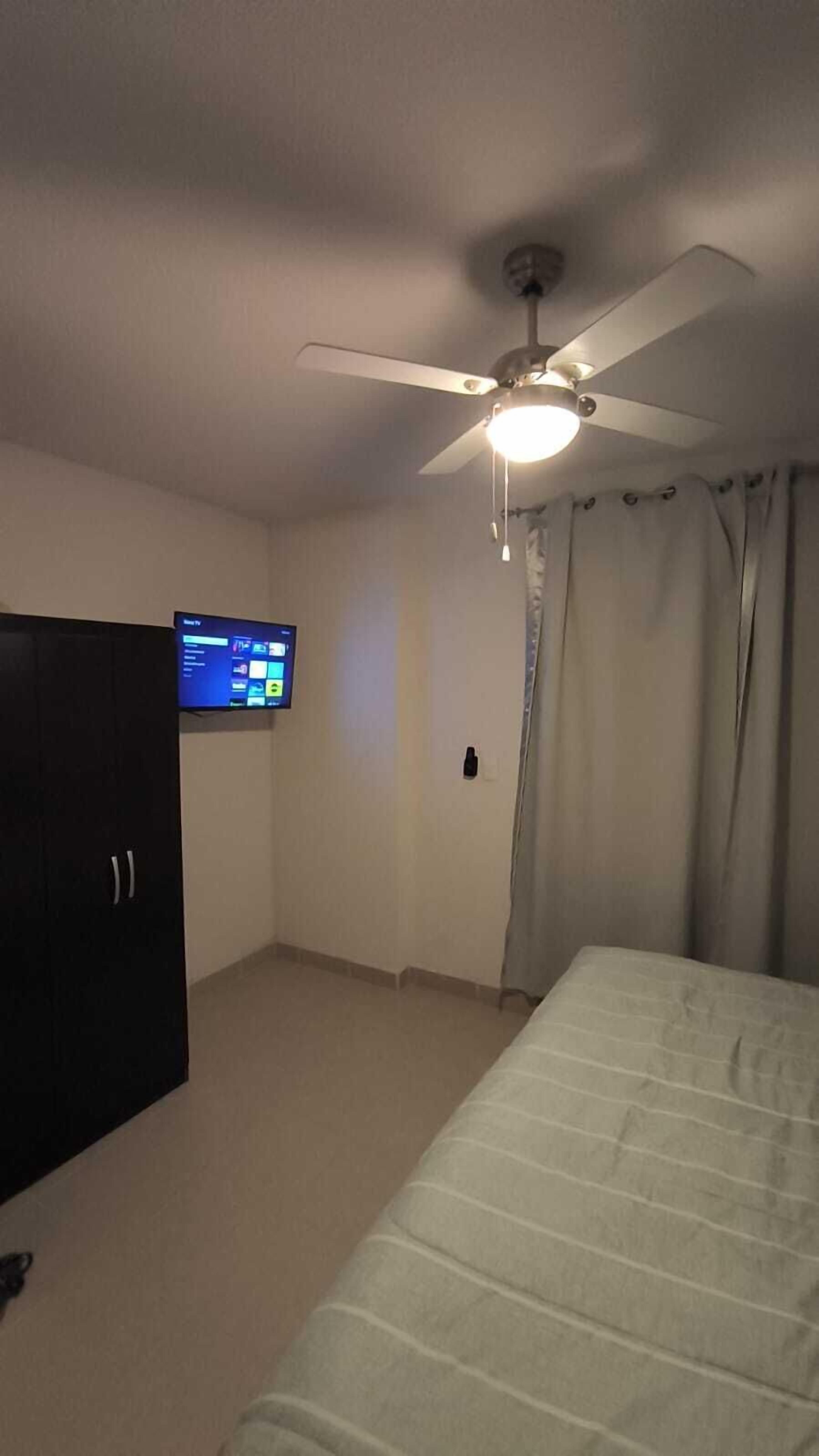 Ein Schlafzimmer mit Bett, Deckenventilator, Fernseher an der Wand und einem dunkelbraunen Kleiderschrank.