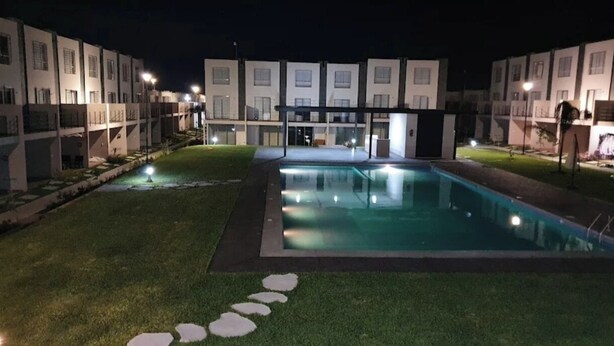 Ein Poolbereich mit klarer Wasserfläche, umgeben von einem gepflegten Rasen und einem gepflasterten Weg, der zu einem Gebäude mit mehreren Fenstern führt.