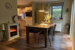 A dining area with a wooden table, chairs, a fireplace, and a clock on the wall. - Cozy Forest Bungalow for 6 (Ewijk)