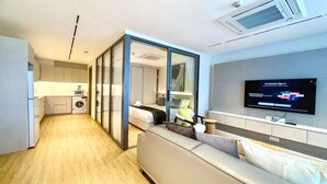 Moderni hotellihuone, jossa on pieni keittiö, sohva, televisio ja liukuovi, joka johtaa kylpyhuoneeseen.