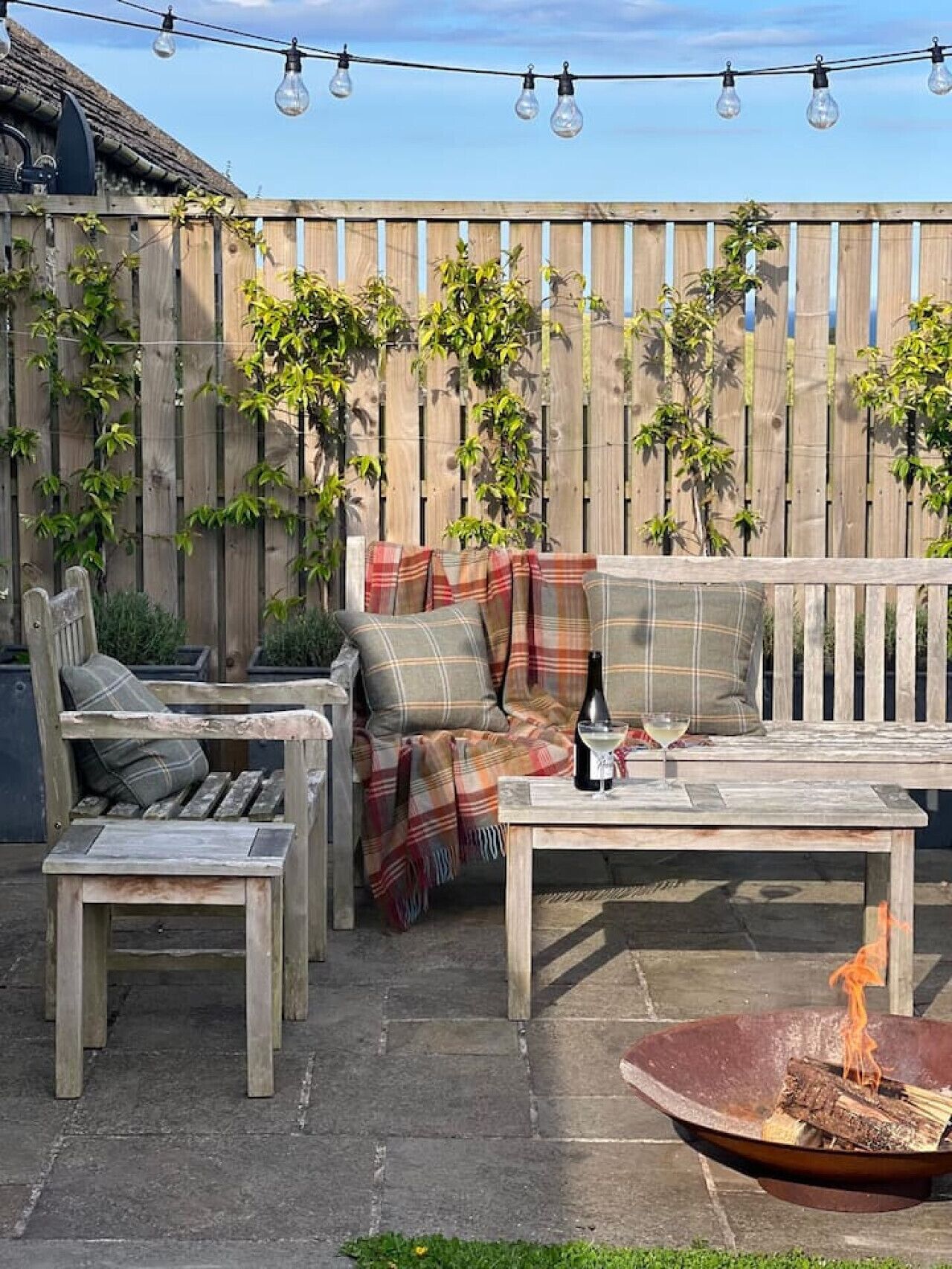 A patio with a wooden bench, a table with a bottle and glass, and a fire pit.
