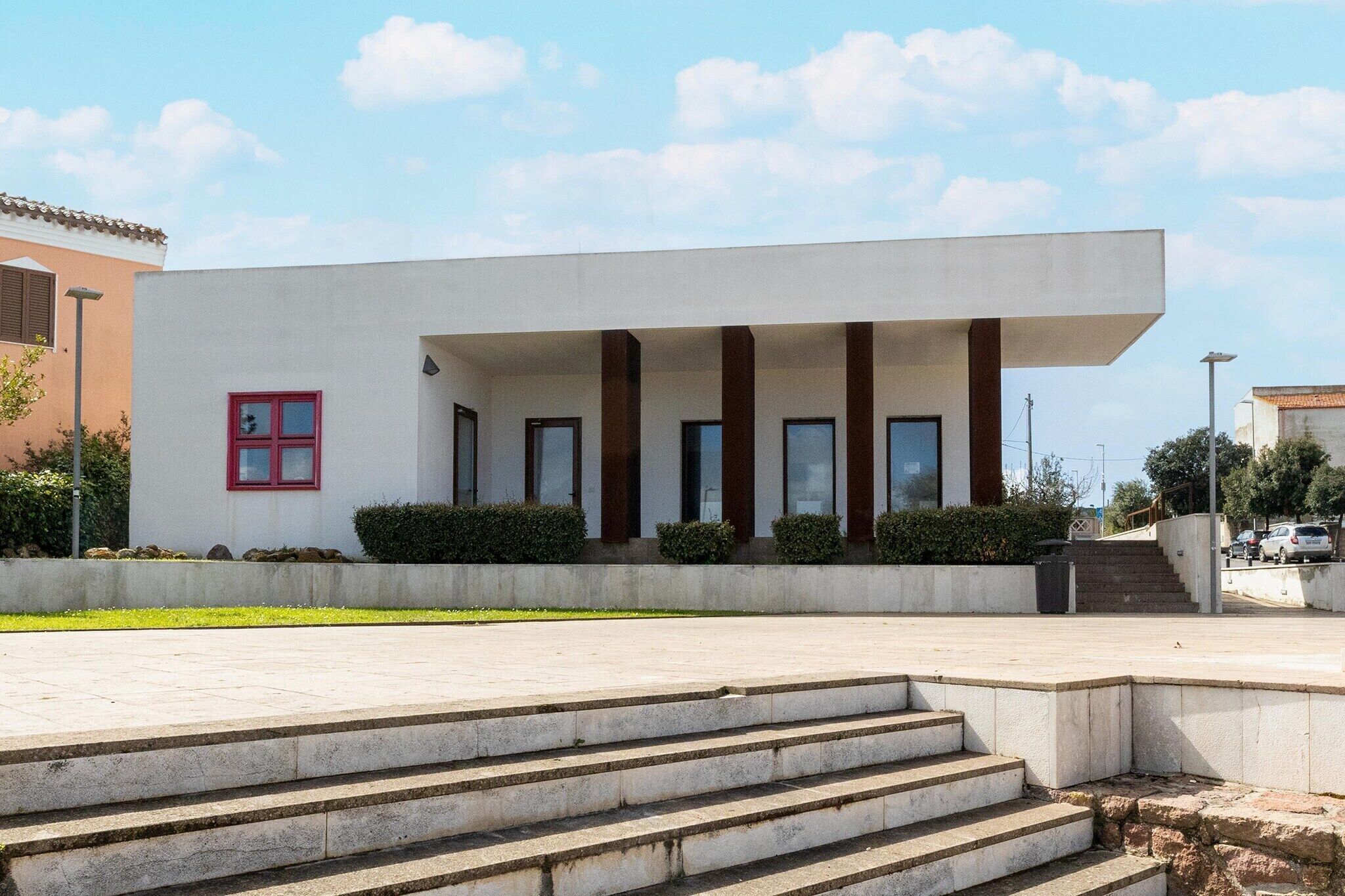 Ein modernes Gebäude mit Flachdach, einer Treppe, die zu einem überdachten Eingang führt, und einem kleinen, rot gefassten Fenster.