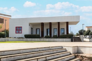 A modern building with a flat roof, a set of stairs leading to a covered entrance, and a small red-framed window. - Apartment with air conditioning (Tergu)
