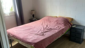 Une chambre avec un lit simple, un plancher en bois, un mur en briques blanches, une table de chevet avec une lampe et une fenêtre avec des rideaux.