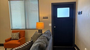 Una sala de estar con sofá, sillón, lámpara, puerta y ventana con persianas.