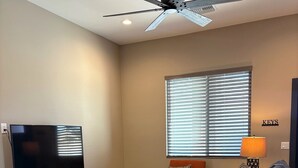Una sala de estar con ventilador de techo, un televisor sobre un mueble de madera, un sofá, un sillón, una alfombra y una ventana con persianas.