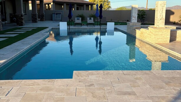 Una piscina con agua azul cristalina, rodeada por un patio con baldosas de piedra, y un edificio con techo de tejas al fondo.