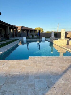 Una piscina con agua azul cristalina, rodeada por un patio con baldosas de piedra, y un edificio con techo de tejas al fondo.