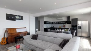 A modern living room with a grey sofa, a glass coffee table, a wooden piano, and a kitchen in the background. - Beautiful modern 4 star apartment with sea view (Dramalj)