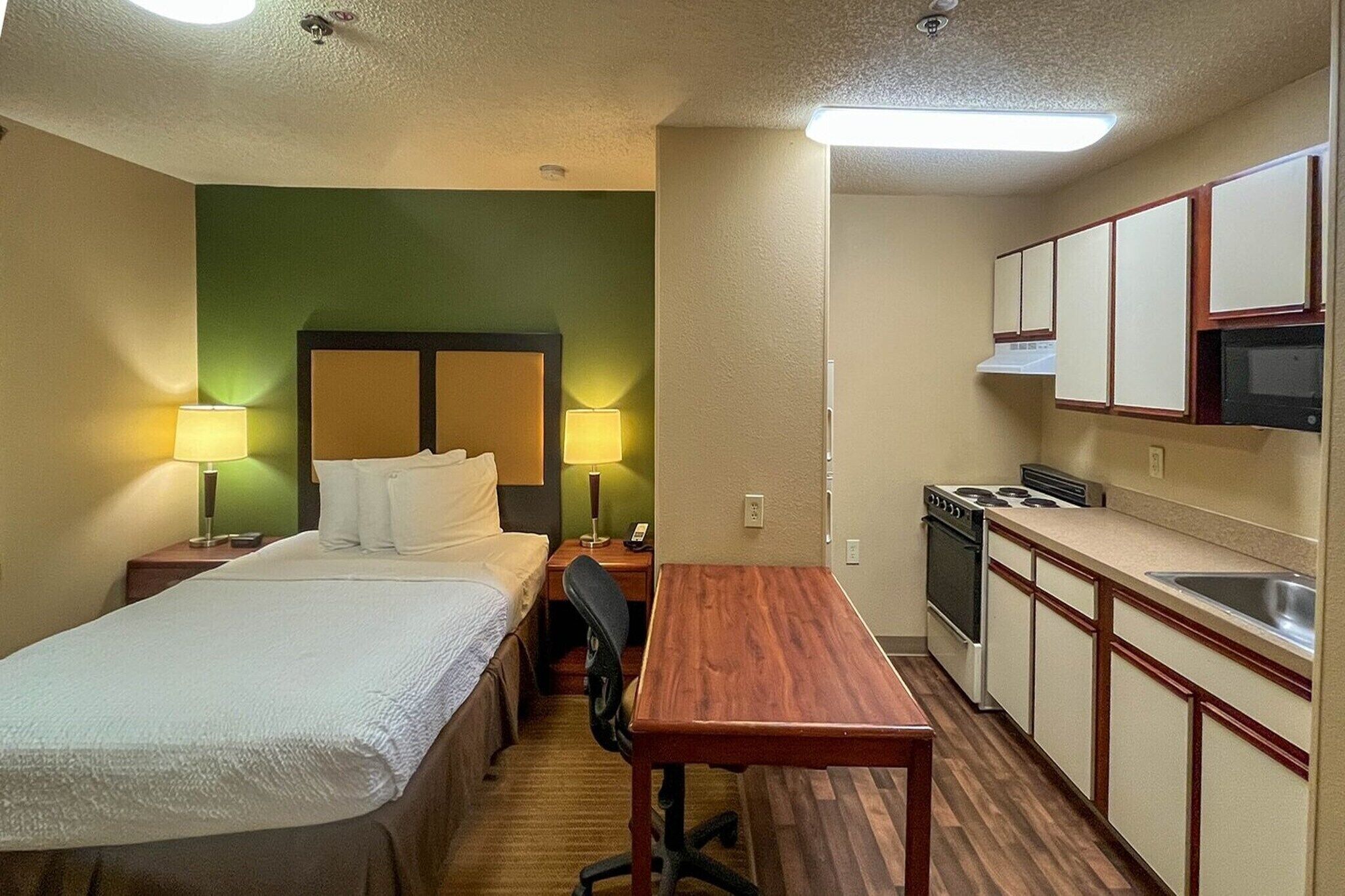 A hotel room with a bed, bedside tables, a desk, a chair, a kitchenette with a stove and microwave, and a green accent wall.