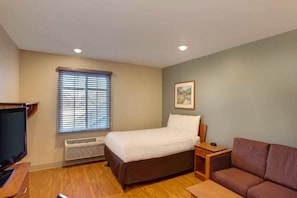A hotel room with a bed, a television, a sofa, a window with blinds, and a framed picture on the wall.