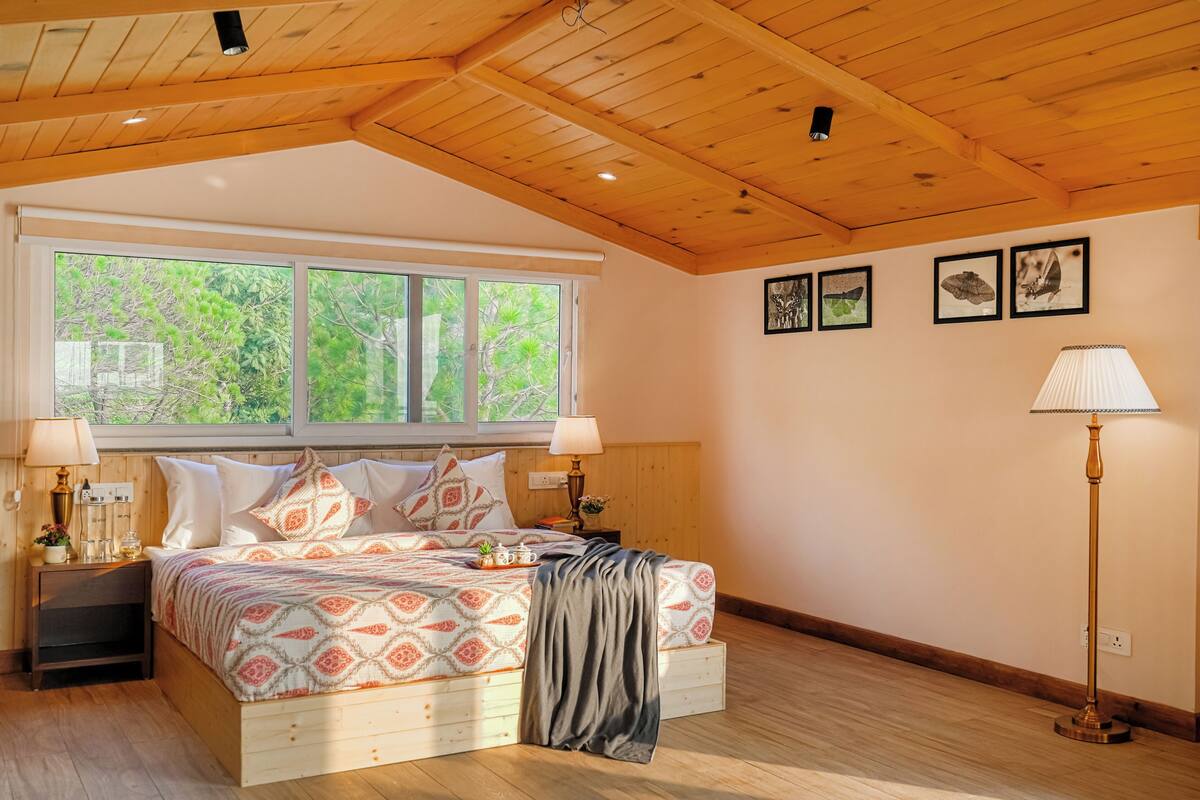 Un dormitorio con techo de madera, una cama grande con ropa de cama estampada, dos lámparas de noche, una mesita de noche y una ventana con vistas a la vegetación.