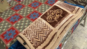 Un lit simple avec une couvre-lit à motifs, un coussin décoratif et une armoire de couleur sombre, dans une pièce aux murs blancs et au sol carrelé.