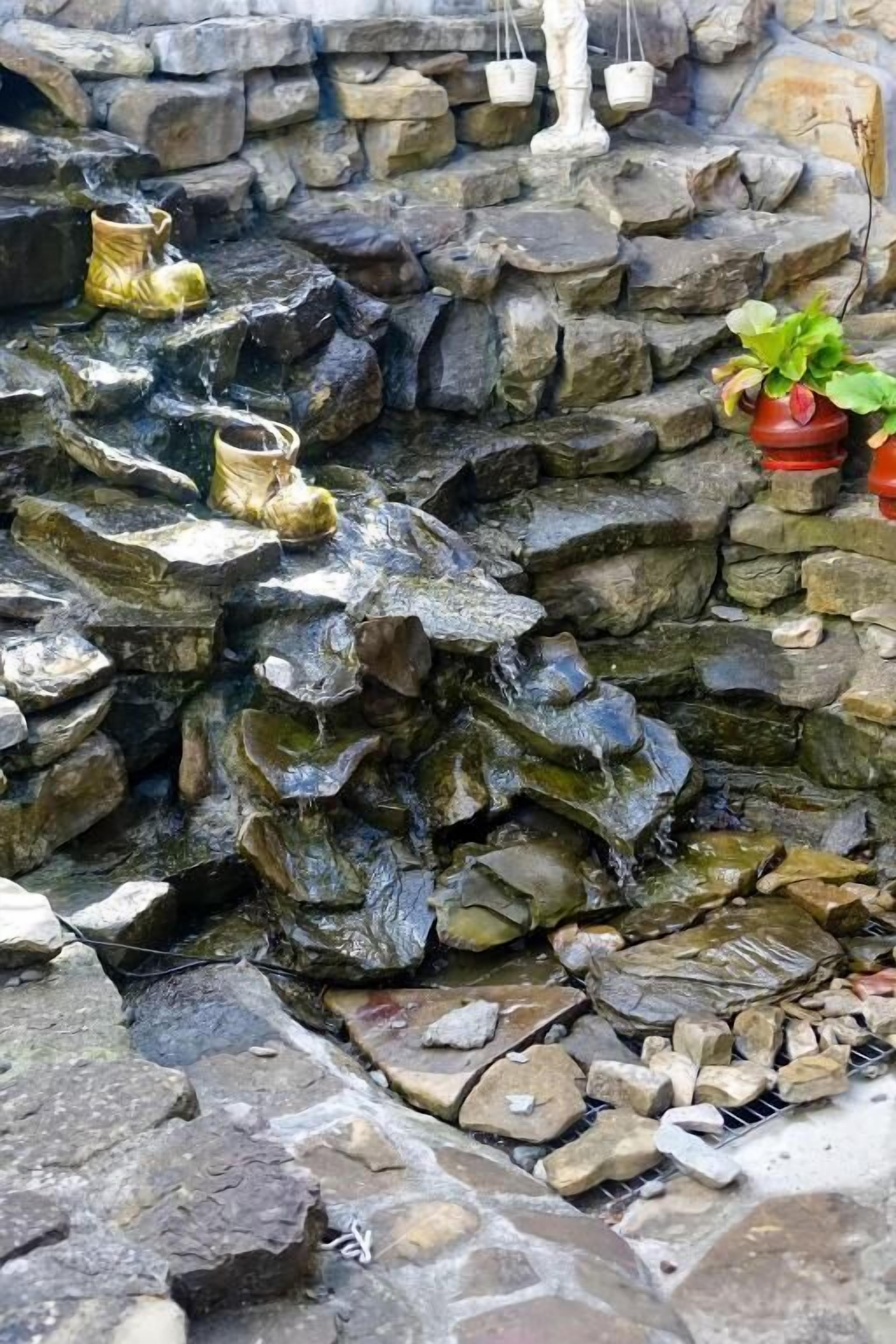 Una fuente de piedra con una pequeña cascada, rodeada de rocas y plantas en macetas.