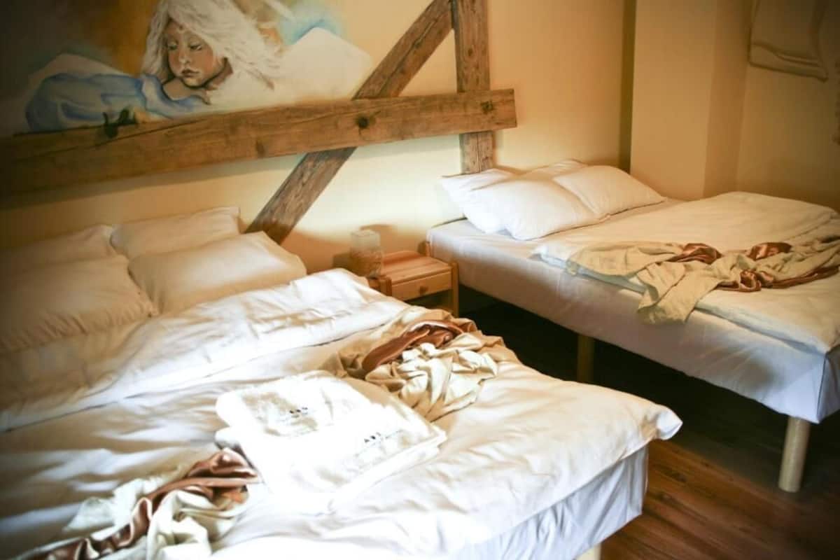 A room with two beds, one with a wooden headboard and the other with a plain headboard. There is a small bedside table with a lamp and a wall-mounted shelf above the beds.