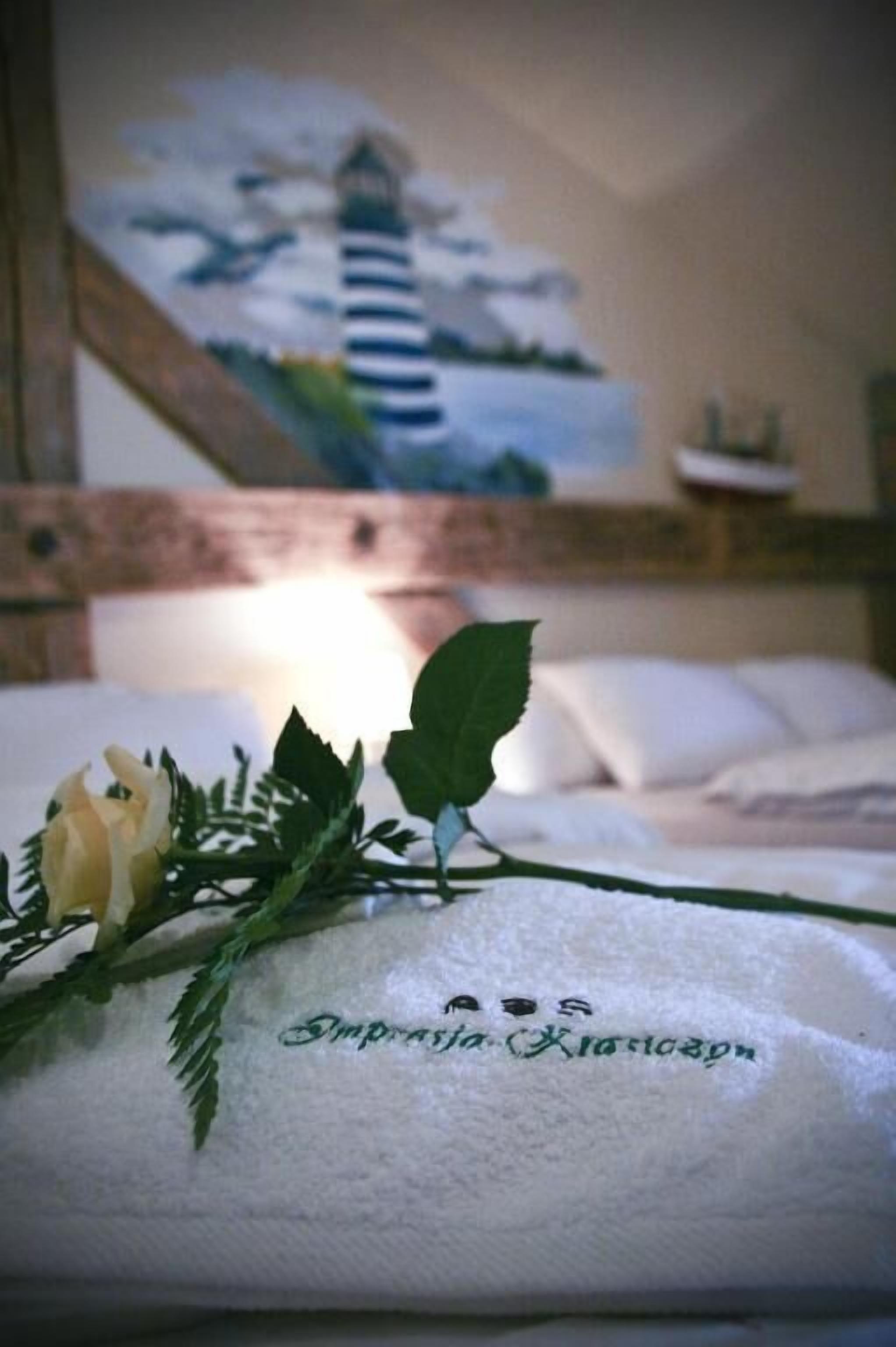 A hotel room with a bed, a painting of a lighthouse, and a towel with a name embroidered on it.