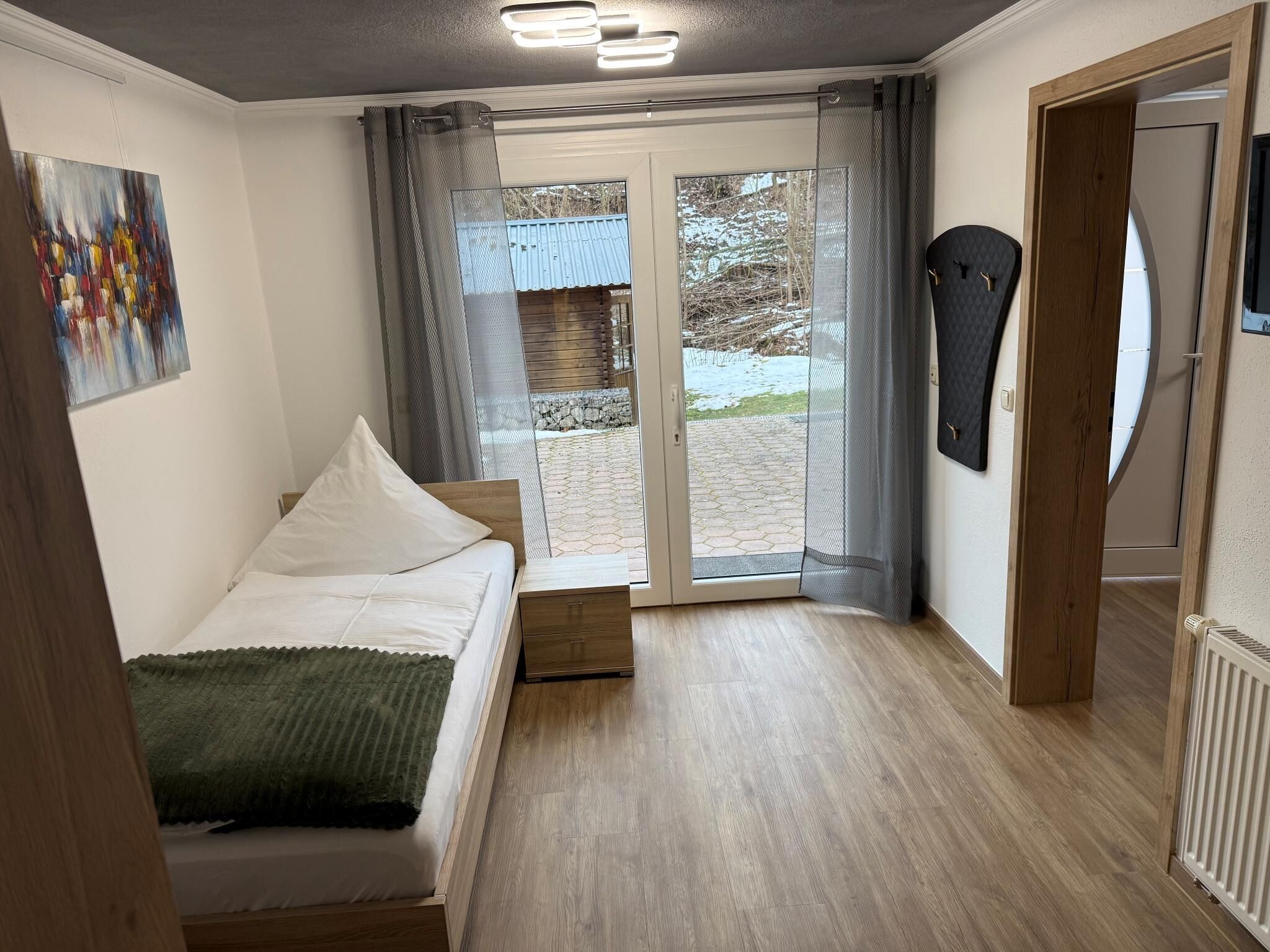 Ein modernes Schlafzimmer mit Bett, einem kleinen Tisch, einem Gemälde und Blick auf einen verschneiten Garten durch eine Glastür.