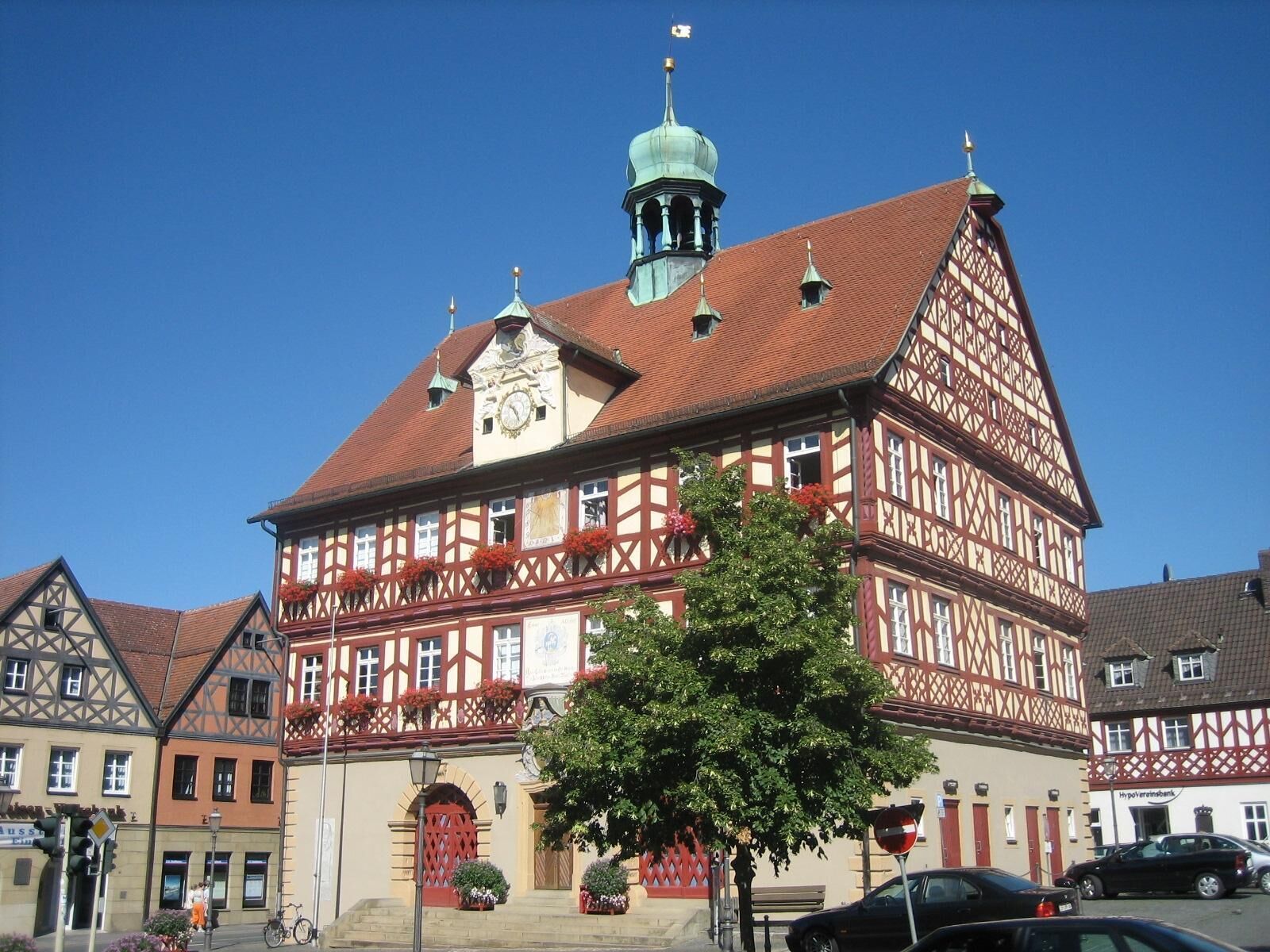 A traditional half-timbered building with a clock tower, a red door, and a tree in front.