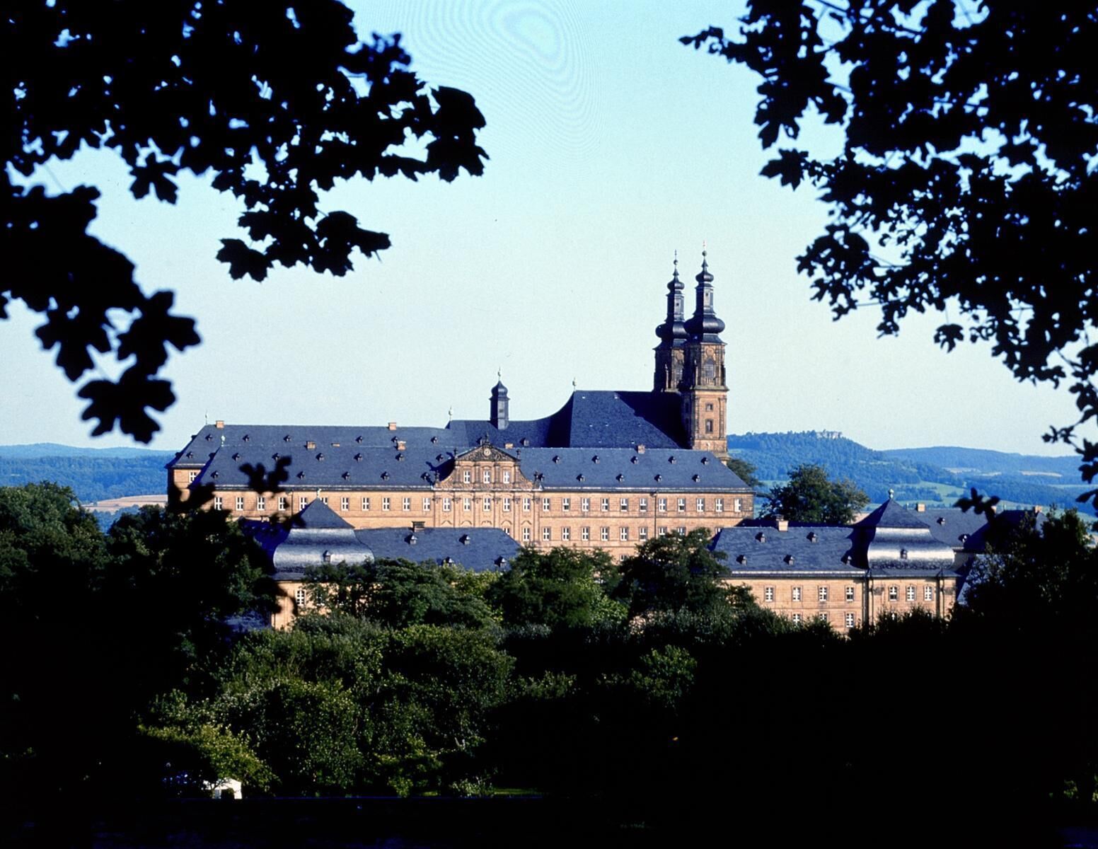 A large historic building with multiple towers and a prominent central tower, surrounded by lush greenery.
