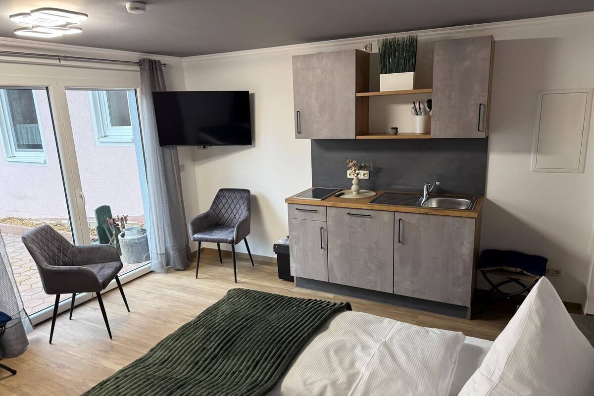 Een moderne kitchenette met een flatscreen-televisie, een zithoek met een stoel, en een bed met een groene deken.
