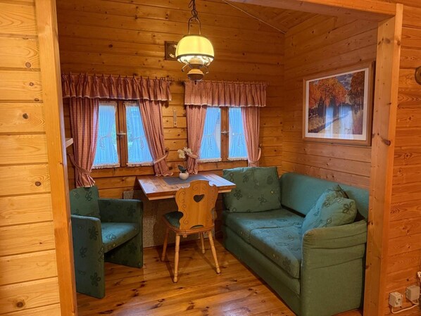 A cozy cabin interior with wooden walls, a dining table, two green upholstered chairs, a wooden desk, a hanging light fixture, and a framed picture on the wall.