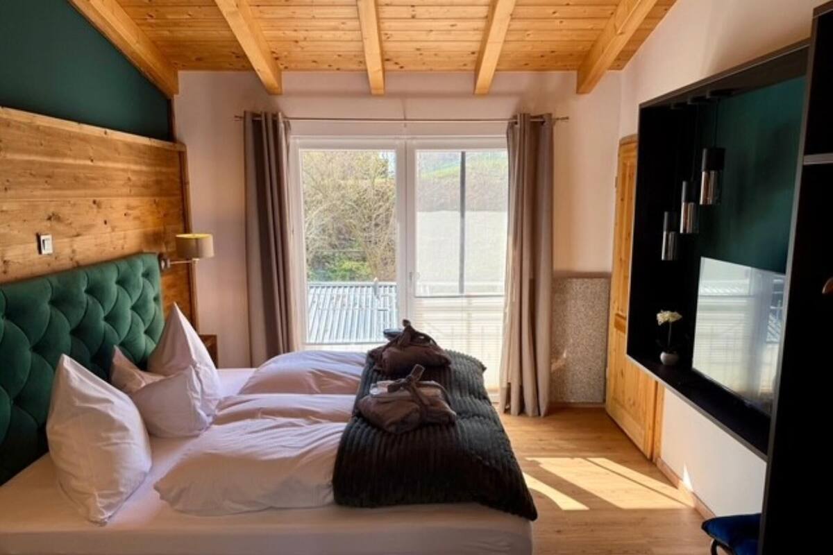 Een slaapkamer met een houten plafond, een groot bed met witte beddengoed, een groen hoofdeinde en uitzicht op het landschap door een raam.