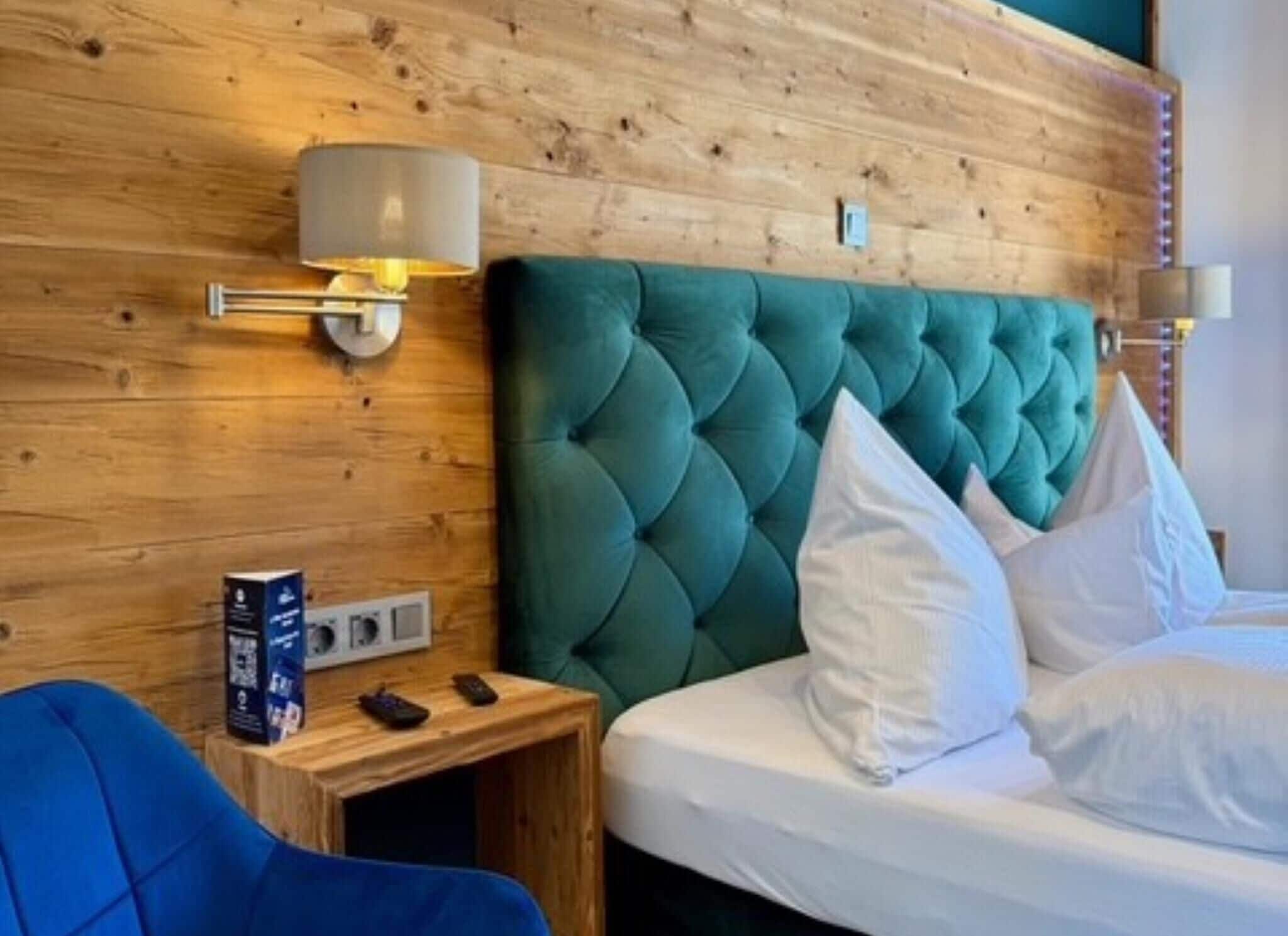 Une chambre d’hôtel avec une tête de lit en bois, une tête de lit capitonnée verte, du linge de lit blanc, une table de chevet avec une lampe et une chaise bleue.