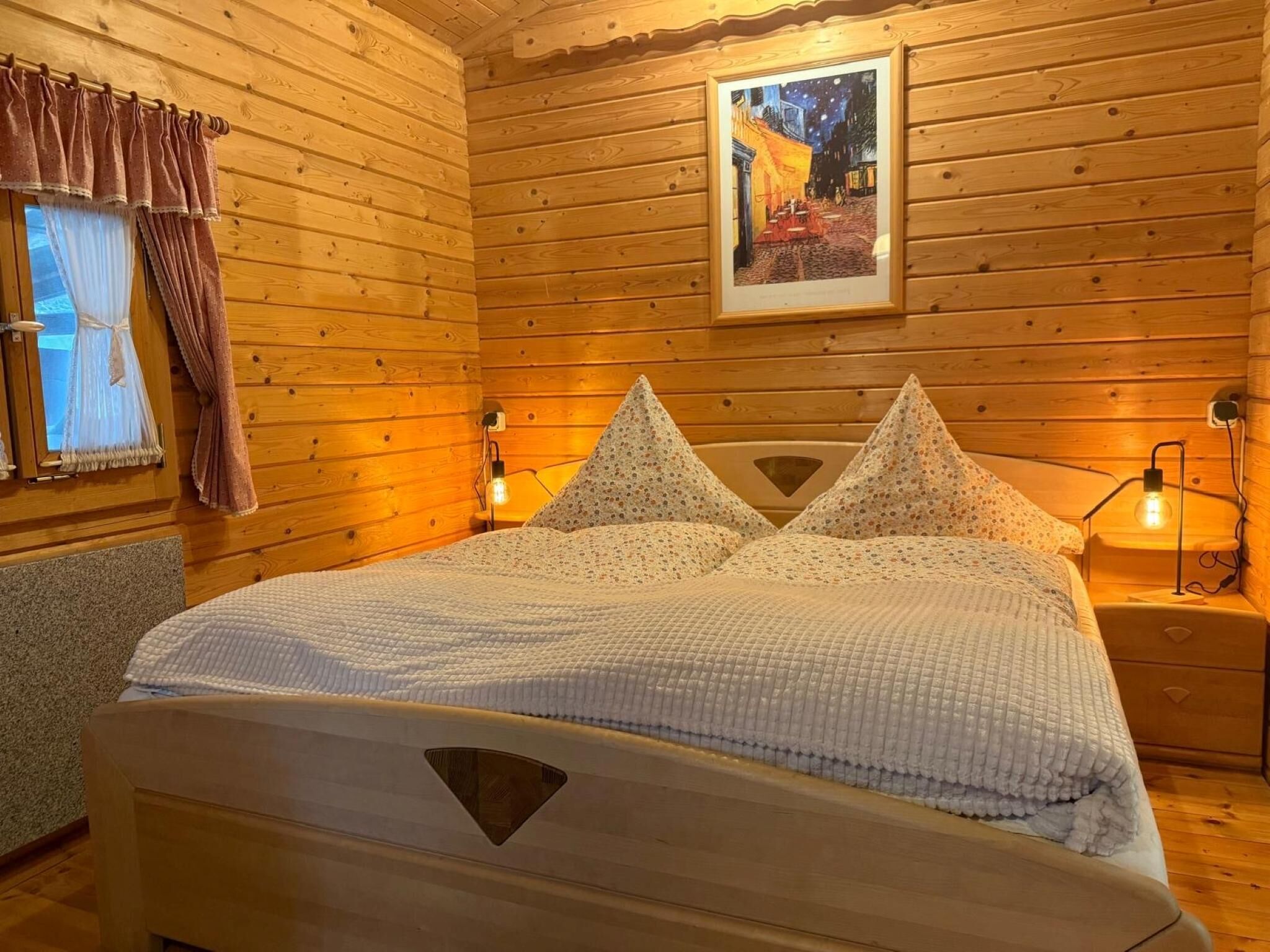 A wooden cabin-style bedroom with a bed, two bedside lamps, a framed painting, and a window with curtains.