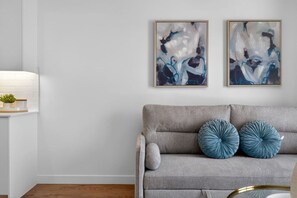 A neutral-toned living room with a grey sofa, two abstract paintings, and a small plant on a white console table. - 2BR | Downtown | AC | Mins to Saddledome (Calgary)