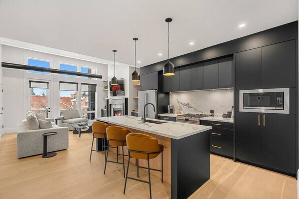 A modern kitchen with a central island, black cabinets, and a large window. - Ember House | Lux 2 Bed | Min to DT (Calgary)