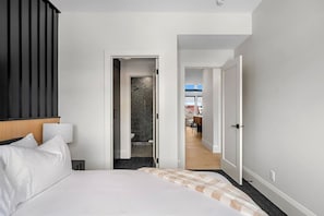 A hotel room with a bed, a nightstand, a bathroom with a shower, and a view of the dining area. - Ember House | Lux 2 Bed | Min to DT (Calgary)