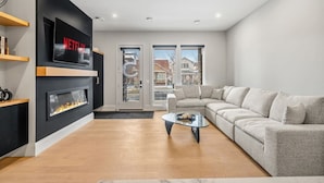 A modern living room with a fireplace, a sectional sofa, a TV mounted on the wall, and a large window. - Onyx Escape | Lux 2 Bed | Garage | Min to DT (Calgary)