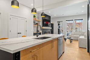 Eine moderne Küche mit Marmorarbeitsplatte, Holzschränken, einem integrierten Geschirrspüler, Pendelleuchten und einem Kamin.