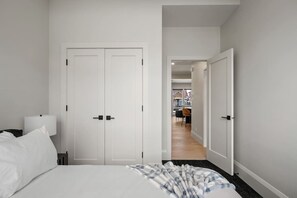 A bedroom with a bed, a nightstand, a lamp, and a closet. There is a hallway leading to another room. - The Platinum Stay | Luxury 2 Bed | Min to DT (Calgary)