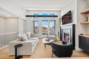 Ein modernes Wohnzimmer mit Sofa, Sessel, Couchtisch, Kamin, Fernseher und großen Fenstern.