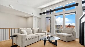 En moderne stue med sofa, lænestol og sofabord, store vinduer med udsigt over byen, og trægulv.