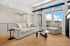 Ein modernes Wohnzimmer mit Sofa, Sessel und Couchtisch, großen Fenstern mit Stadtblick und Holzboden.