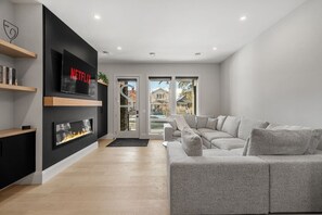 Ein modernes Wohnzimmer mit grauem Sofa, Kamin, Wand-Fernseher und Blick ins Freie.
